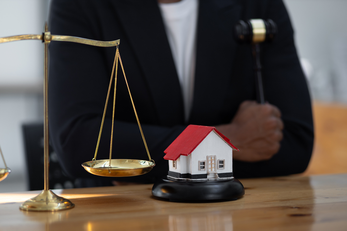 A lawyer holding a gavel sitting behind a desk with a model home and justice scales